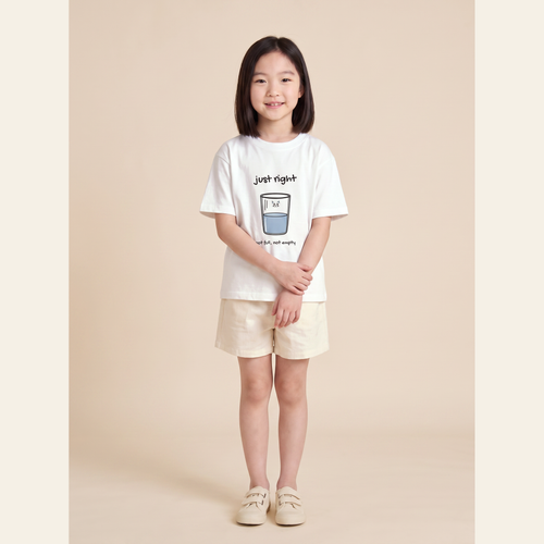 Girl standing in the White kids tee on a warm cream studio background.