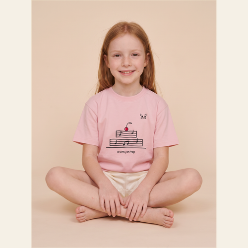 Sitting cross-legged in the Pink kids tee, cream studio.